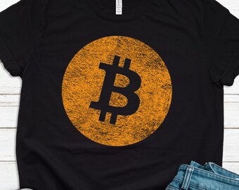 May include: Black t-shirt with a large orange circle featuring a black Bitcoin symbol. The shirt is laid flat on a white wooden surface with a pair of white sneakers and blue jeans.