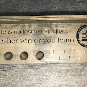 Op de afbeelding: Houten jiu-jitsu display met de tekst "There is no losing in jiu jitsu. You either win or you learn." Voorzien van een Third Generation Brazilian Jiu-Jitsu logo, houten pinnen en haken.