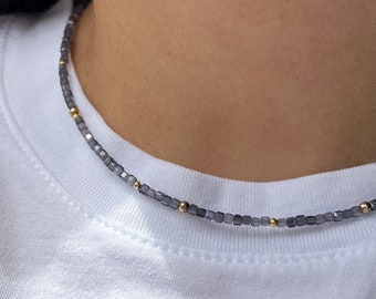 May include: A delicate necklace featuring small, dark gray square beads interspersed with tiny gold-colored beads. The necklace is worn against a white t-shirt, creating a simple yet elegant look. The necklace appears to be a choker style.