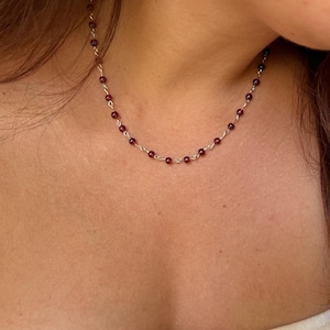 Op de afbeelding: Een delicate ketting met kleine, ronde, dieprode granaatkralen die gelijkmatig verdeeld zijn over een dunne, heldere, zilverkleurige metalen ketting. De schakels van de ketting zijn gedraaid en verbonden met elke individuele kraal.