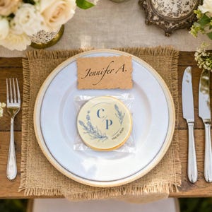 May include: A table setting with a white plate, silverware, and a burlap placemat. A cookie with a floral design and the initials "C P" is on the plate. A name card reads "Jennifer A." Floral arrangements and candles complete the elegant look.