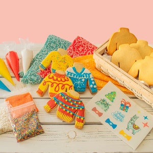 May include: A cookie decorating kit with a variety of colorful icing tubes, sprinkles, and sugar. The kit includes four undecorated sugar cookies shaped like sweaters, and four decorated sweater cookies. The kit also includes a sheet of stickers with holiday-themed designs.