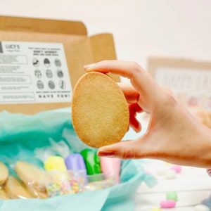 May include: A box filled with cookie decorating supplies, including sugar cookies shaped like Easter eggs, colorful icing, and sprinkles. A hand holds up a plain sugar cookie shaped like an egg.
