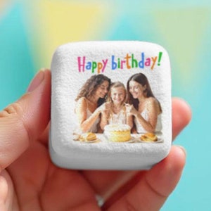 May include: White square stone with a photo of three people celebrating a birthday. The photo shows a girl blowing out candles on a cake while two other people smile and look at her. The text "Happy birthday!" is printed in rainbow colors above the photo.