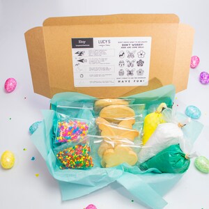 Puede incluir: Una caja de cart&oacute;n marr&oacute;n con un forro de papel de seda azul y blanco. La caja contiene un kit de decoraci&oacute;n de galletas con galletas de az&uacute;car, chispas y glaseado. La caja est&aacute; etiquetada como "Lucy's" y tiene instrucciones para decorar las galletas. La caja est&aacute; rodeada de huevos de Pascua coloridos.