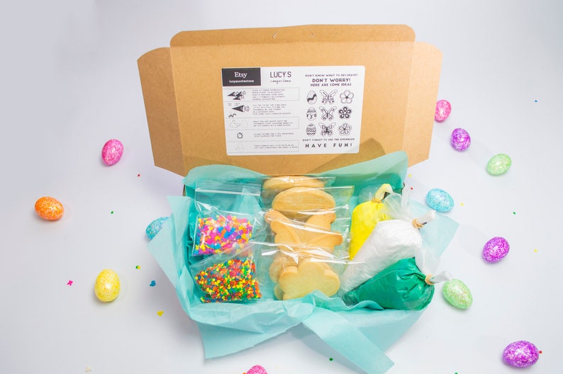 Puede incluir: Una caja de cart&oacute;n marr&oacute;n con un forro de papel de seda azul y blanco. La caja contiene un kit de decoraci&oacute;n de galletas con galletas de az&uacute;car, chispas y glaseado. La caja est&aacute; etiquetada como "Lucy's" y tiene instrucciones para decorar las galletas. La caja est&aacute; rodeada de huevos de Pascua coloridos.