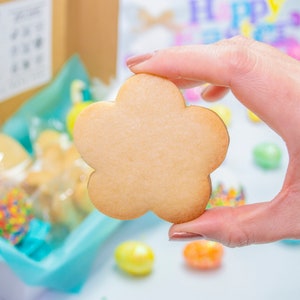 Puede incluir: Una galleta de az&uacute;car simple, de color marr&oacute;n claro, con forma de flor. La galleta est&aacute; siendo sostenida por una mano.