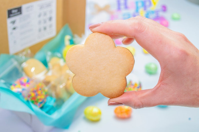 Puede incluir: Una galleta de az&uacute;car simple, de color marr&oacute;n claro, con forma de flor. La galleta est&aacute; siendo sostenida por una mano.