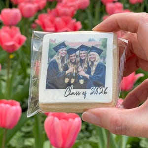 May include: A square cookie with a photo of four graduates in caps and gowns, holding champagne bottles, with "Class of 2026" written on it. The cookie is held in a clear plastic wrapper, with a background of pink tulips.