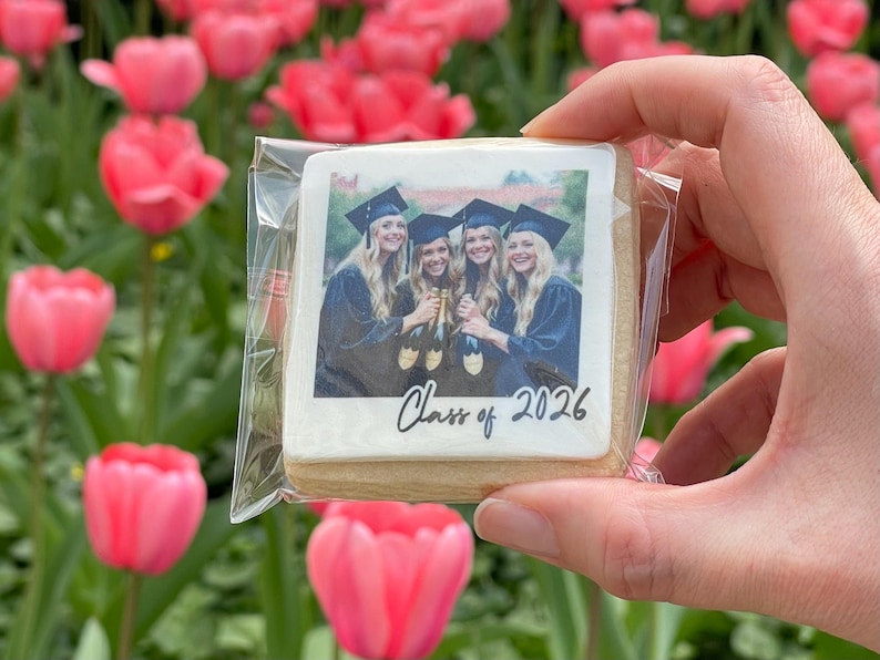 May include: A square cookie with a photo of four graduates in caps and gowns, holding champagne bottles, with "Class of 2026" written on it. The cookie is held in a clear plastic wrapper, with a background of pink tulips.