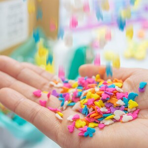 Puede incluir: Una mano sosteniendo una variedad de espolvorear coloridos en forma de mariposas y corazones. Los espolvorear son una mezcla de rosa, azul, amarillo, naranja y blanco.
