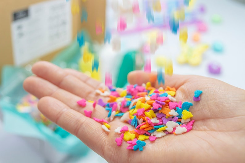 Puede incluir: Una mano sosteniendo una variedad de espolvorear coloridos en forma de mariposas y corazones. Los espolvorear son una mezcla de rosa, azul, amarillo, naranja y blanco.