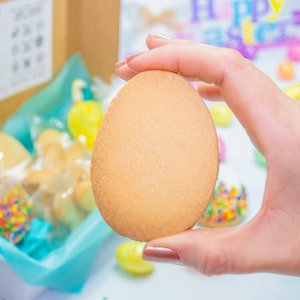 Puede incluir: Una mano sosteniendo una galleta grande, simple, de forma ovalada. La galleta es de color marr&oacute;n claro y tiene una textura ligeramente rugosa. La galleta est&aacute; rodeada de otras galletas y dulces en una caja de regalo.