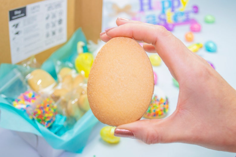 Puede incluir: Una mano sosteniendo una galleta grande, simple, de forma ovalada. La galleta es de color marr&oacute;n claro y tiene una textura ligeramente rugosa. La galleta est&aacute; rodeada de otras galletas y dulces en una caja de regalo.