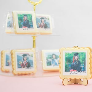 May include: Graduation-themed cookies with edible photo prints. The cookies are square-shaped with scalloped edges and feature a photo of a graduate in a cap and gown. The text "Just Graduated" is printed on the photo. The cookies are displayed on a tiered stand.