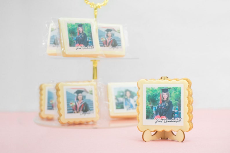 May include: Graduation-themed cookies with edible photo prints. The cookies are square-shaped with scalloped edges and feature a photo of a graduate in a cap and gown. The text "Just Graduated" is printed on the photo. The cookies are displayed on a tiered stand.