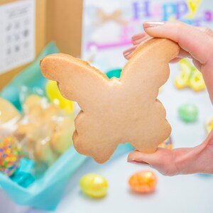 Puede incluir: Una mano sostiene una galleta de az&uacute;car simple de color marr&oacute;n claro con forma de mariposa. La galleta est&aacute; enfocada, mientras que el fondo est&aacute; desenfocado y muestra una caja de galletas, chispas y huevos de Pascua de colores pastel.