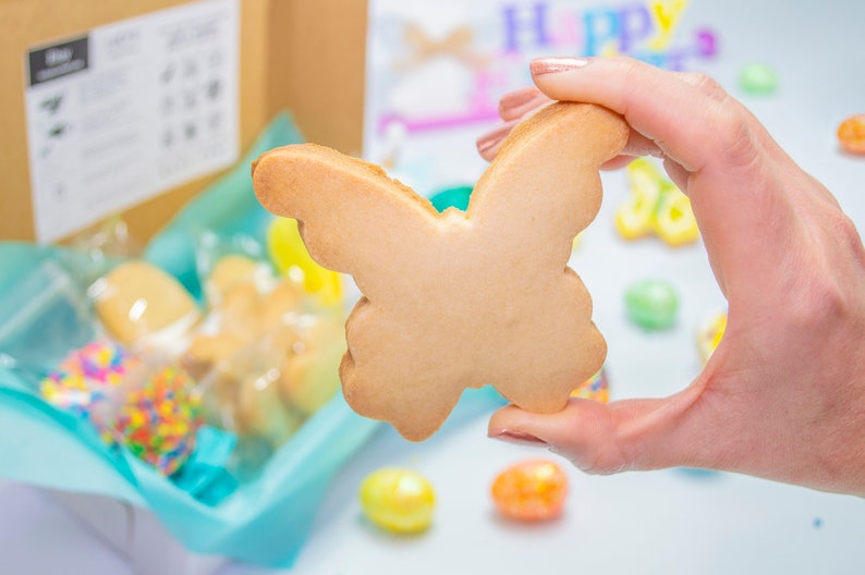 Puede incluir: Una mano sostiene una galleta de az&uacute;car simple de color marr&oacute;n claro con forma de mariposa. La galleta est&aacute; enfocada, mientras que el fondo est&aacute; desenfocado y muestra una caja de galletas, chispas y huevos de Pascua de colores pastel.