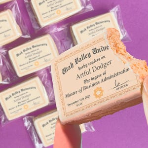 May include: A close-up of a cookie shaped like a diploma, with a bite taken out of it. The cookie has text that reads "Utah Valley Unive" and "Master of Business Administration." Other cookies in clear packaging are in the background on a purple surface.