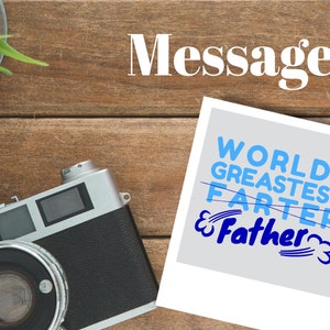 May include: A white Polaroid photo with a blue and white text overlay that says "Worlds Greatest Farter" with the word "Farter" crossed out and "Father" written in below it. The photo is on a wooden surface with a vintage camera.