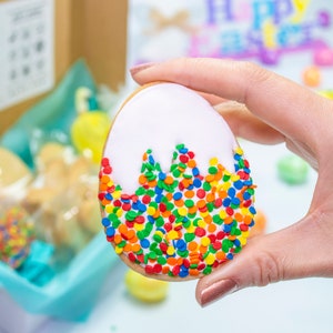 Puede incluir: Una galleta de az&uacute;car blanca y amarilla con forma de huevo, decorada con chispas de colores. La galleta est&aacute; siendo sostenida por una mano.