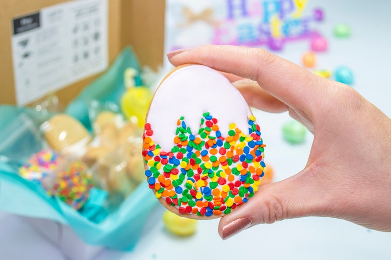 Puede incluir: Una galleta de az&uacute;car blanca y amarilla con forma de huevo, decorada con chispas de colores. La galleta est&aacute; siendo sostenida por una mano.