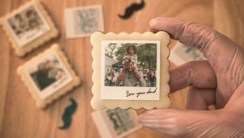 May include: A white biscuit with a scalloped edge, decorated with a photo of a father and child. The photo shows a girl sitting on her father's shoulders. The biscuit has the text "Love you dad" written on it.