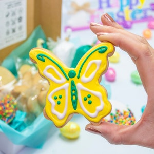 Puede incluir: Una galleta con forma de mariposa amarilla con glaseado verde y blanco. La galleta est&aacute; siendo sostenida por una mano.
