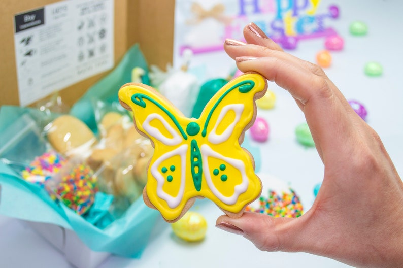 Puede incluir: Una galleta con forma de mariposa amarilla con glaseado verde y blanco. La galleta est&aacute; siendo sostenida por una mano.