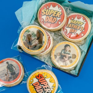 May include: A box of decorated cookies with images and text. Some cookies say "Super Dad" in a comic book style, while others have family photos and "Happy Father's Day" on them. The cookies are round and individually wrapped.