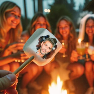 May include: A marshmallow on a stick with an image of George Clooney, held in front of a campfire. Several people are gathered around the fire, smiling and holding drinks. The scene is lit by the firelight.