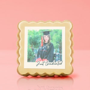 May include: A graduation-themed cookie with a photo of a graduate in a cap and gown. The photo is framed with a light-colored icing border and the words "Just Graduated" are printed on the photo. The cookie has a scalloped edge.