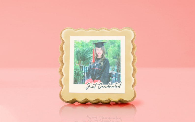 May include: A graduation-themed cookie with a photo of a graduate in a cap and gown. The photo is framed with a light-colored icing border and the words "Just Graduated" are printed on the photo. The cookie has a scalloped edge.