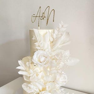 Peut inclure: Un gâteau de mariage blanc à deux niveaux décoré de fleurs et de verdure blanches. Un dessus doré affiche les lettres "A&N". Le gâteau est orné de grandes roses ouvertes et de divers éléments floraux, créant un design romantique et élégant.