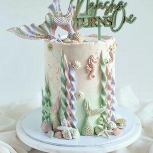 Op de afbeelding: Een verjaardagstaart met zeemeermint thema in pastelkleuren. De taart is versierd met zeemeerminstaarten, zeepaardjes en schelpen. De taarttopper luidt "Natasha Turns One". De taart staat op een witte taartstandaard.