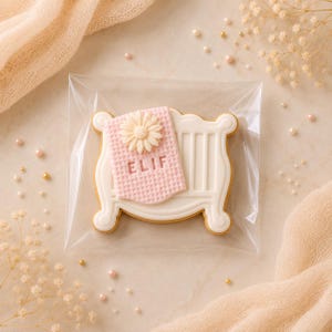 Op de afbeelding: Een koekje in de vorm van een wit babywiegje met een roze, getextureerde deken eroverheen gedrapeerd. De deken is voorzien van een witte madeliefbloem en de naam ELIF in hoofdletters. Het koekje zit verpakt in een doorzichtig plastic zakje.