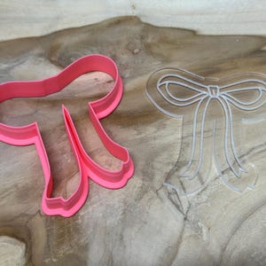 Puede incluir: Un cortador de galletas rosa en forma de lazo y una plantilla transparente en forma de lazo sobre una superficie de madera. El cortador de galletas es de plástico rosa y tiene un diseño detallado de lazo. La plantilla es transparente.