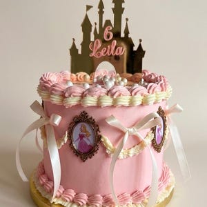 May include: A pink frosted cake decorated with white piped borders, cream-colored ribbons, and oval frames featuring princess illustrations. A gold-colored acrylic cake topper shaped like a castle displays the number 6 and the name Leila.