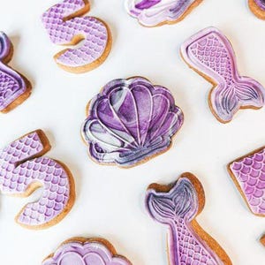 May include: Overhead view of mermaid-themed biscuits. The biscuits are decorated with purple and white icing, featuring seashell, mermaid tail, and number 5 shapes. The biscuits are arranged on a white surface.