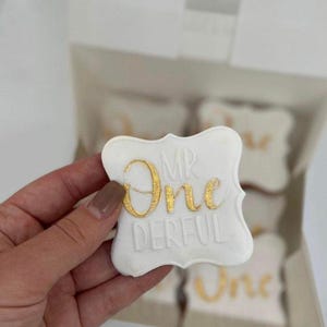 May include: A white cookie with gold lettering that says "MR One DERFUL". The cookie is held in a hand and is in front of a box of similar cookies. The cookies are likely for a first birthday celebration.