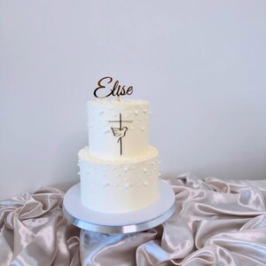 Può includere: Una torta bianca a due piani con decorazioni a forma di perla. Un topper in legno con la scritta "Elise" sopra un disegno a croce e colomba. La torta è su un supporto bianco, esposta su un tessuto di raso.
