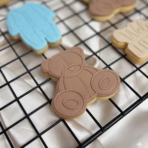 Op de afbeelding: Een verzameling versierde koekjes, waaronder een bruine teddybeer, een blauwe onesie en een koekje met de tekst "Welcome Little One". De koekjes zijn gerangschikt op een zwart draadrek.