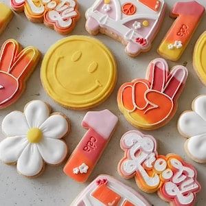 Op de afbeelding: Een verzameling versierde suikerkoekjes met een retro thema. Ontwerpen omvatten een gele smiley, vredestekens, bloemen en een peace-thema busje. De koekjes zijn versierd met witte, oranje, roze en gele glazuur.