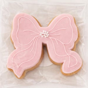 Op de afbeelding: Een roze koekje in de vorm van een strik met witte glazuurdetails en een cluster van witte sprinkles in het midden. De koek heeft een lichtbruine, gebakken rand en wordt gepresenteerd op een doorzichtig, gekreukt oppervlak.