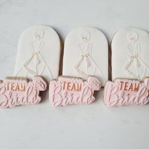 Op de afbeelding: Drie versierde suikerkoekjes. Elk koekje heeft een witte boogvormige bovenkant met een bruidsillustratie en een roze koekje eronder met de tekst "Team Bride" in gouden letters. De koekjes liggen op een wit oppervlak.