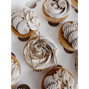 Pode incluir: Uma dúzia de cupcakes numa caixa branca, cada um coberto com glacê cinza e branco. Alguns cupcakes têm coberturas douradas que dizem "Oh Baby". As raspas adicionam detalhes à cobertura. Os cupcakes estão numa caixa de padaria branca.