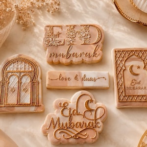 May include: A collection of four decorated cookies with a marble-like beige and white finish. One cookie features the text EID MUBARAK with floral details, another shows an arched window, one is a rectangular bar with love & duas, and one is a cloud-shaped cookie with EID MUBARAK and a crescent moon.