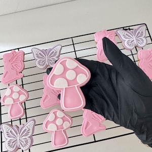 Puede incluir: Galletas rosas y blancas glaseadas con forma de setas, mariposas y hadas dispuestas sobre una rejilla negra. Las galletas están decoradas con detalles de glaseado blanco. Una mano enguantada de negro es parcialmente visible.