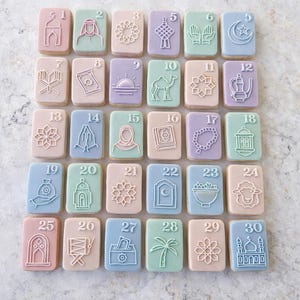 Peut inclure: Une sélection de 30 biscuits rectangulaires, chacun avec un motif en relief et un numéro différent de 1 à 30. Les biscuits sont de couleurs pastel, notamment rose, bleu, vert et violet, avec des détails blancs. Les motifs incluent des symboles et objets religieux.
