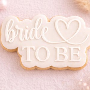 Op de afbeelding: Een versierde suikerkoek met de woorden "bride TO BE" in witte glazuur. De koek heeft de vorm van een wolk en heeft een hartvormig ontwerp. De koek ligt op een roze ondergrond.